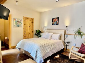 Een slaapkamer met een bed, een houten deur, een schilderij, een wandlamp, een kleine tafel met een plant en een stoel.