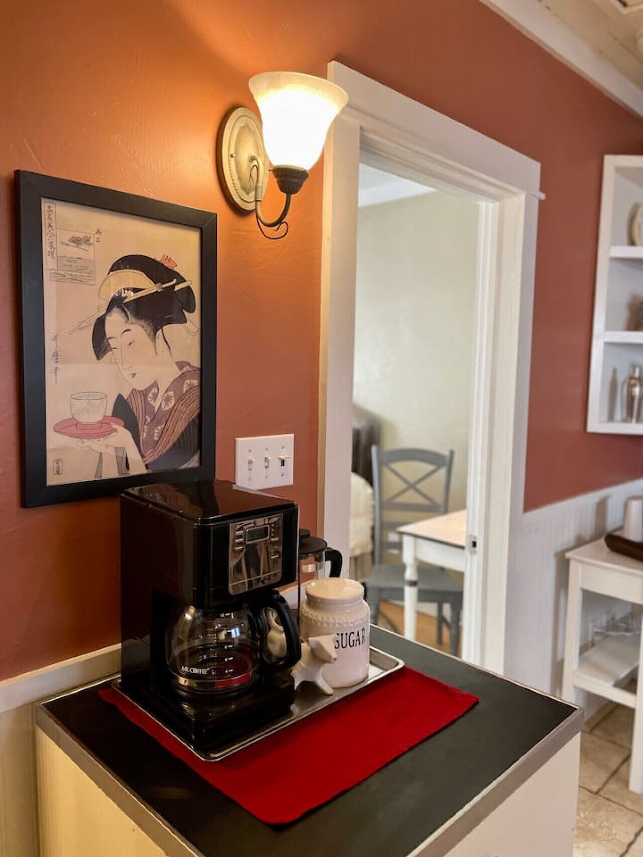 Une machine à café posée sur un tapis rouge, à côté d’un récipient à sucre, un tableau encadré accroché au mur et une porte-fenêtre donnant sur un coin repas.