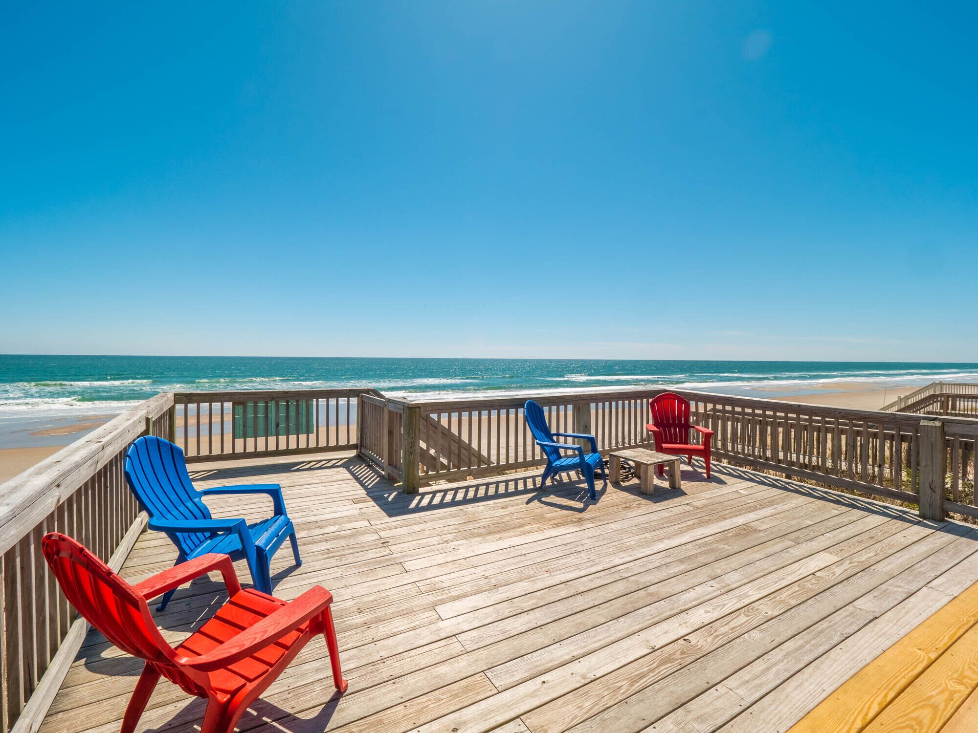 Une terrasse en bois avec des fauteuils Adirondack bleus et rouges donnant sur l’océan.