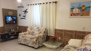 A living room with a floral sofa, a glass coffee table, a television on a wooden stand, and a painting on the wall. - Residencial Dois Amores 2 (Florianópolis)