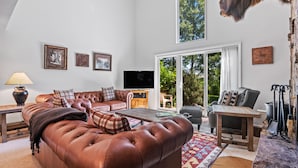 A spacious living room with a brown leather sofa, a flat-screen TV, and a large window providing a view of the outdoors. - RMR: Four Seasons II Unit 1 Condo in Teton Village (Teton Village)