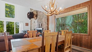 A dining area with a wooden table and chairs, a stone fireplace, and a large window with a view of trees. - RMR: Four Seasons II Unit 1 Condo in Teton Village (Teton Village)