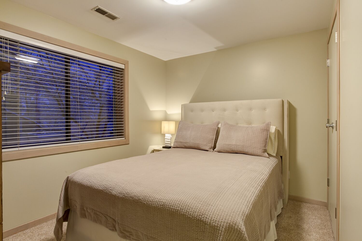 Ein Schlafzimmer mit einem Bett, einem Nachttisch, einer Lampe und einem Fenster mit Jalousien.