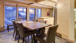 Een eetgedeelte met een houten tafel en stoelen, een keuken met een koelkast, en grote ramen met uitzicht op buiten.