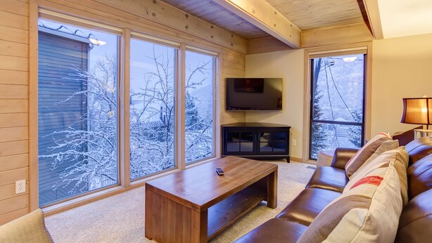 Een gezellige woonkamer met een houten bank, een aan de muur bevestigde televisie en grote ramen met uitzicht op besneeuwde bomen.