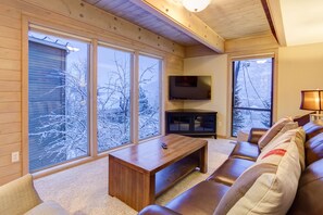 A cozy living room with a wooden bench, a TV mounted on the wall, and large windows offering a view of snow-covered trees. - Right in the heart of Old Town, this fantastic cabin is one of the best rentals in all of Park City! (Park City)