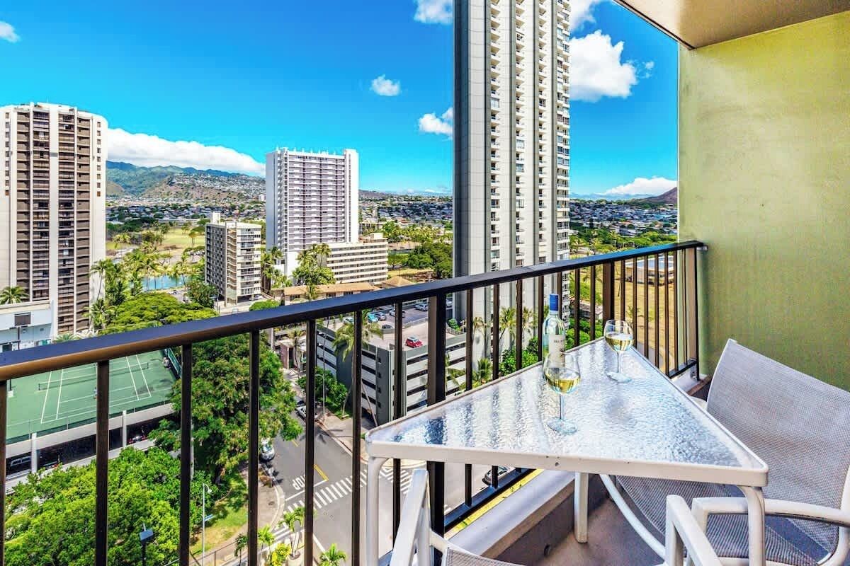 A balcony with a table and chairs, overlooking a cityscape with high-rise buildings and a tennis court.
