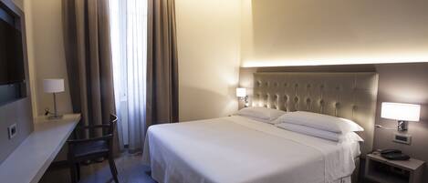 A hotel room with a large bed, a desk, a chair, a nightstand, and a TV.