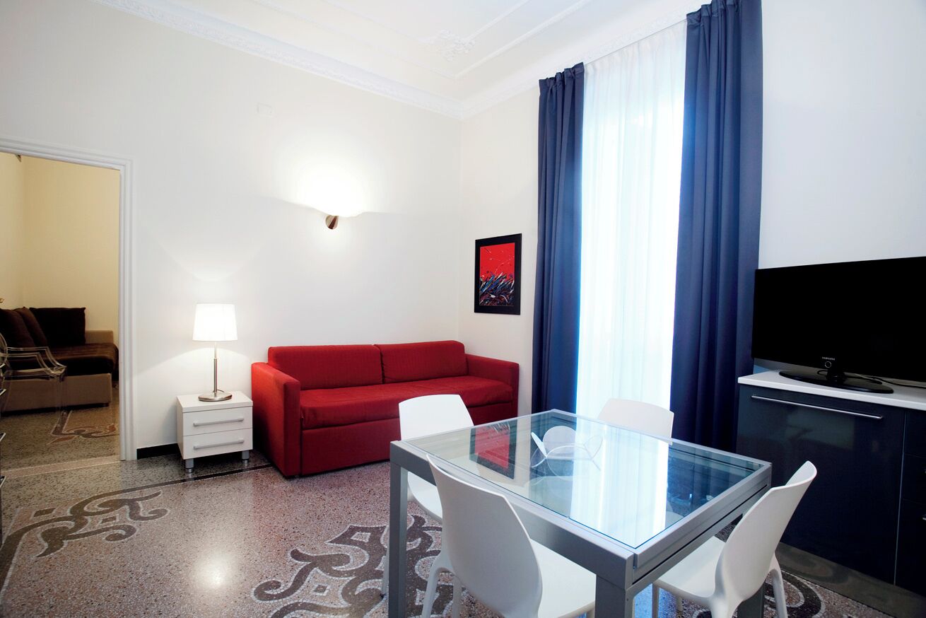 Habitación de hotel con mesa de vidrio, sillas blancas, sofá rojo, una pequeña mesita de noche blanca con lámpara, televisor de pantalla plana y un amplio ventanal con cortinas azules.