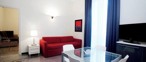 Habitación de hotel con mesa de vidrio, sillas blancas, sofá rojo, una pequeña mesita de noche blanca con lámpara, televisor de pantalla plana y un amplio ventanal con cortinas azules.