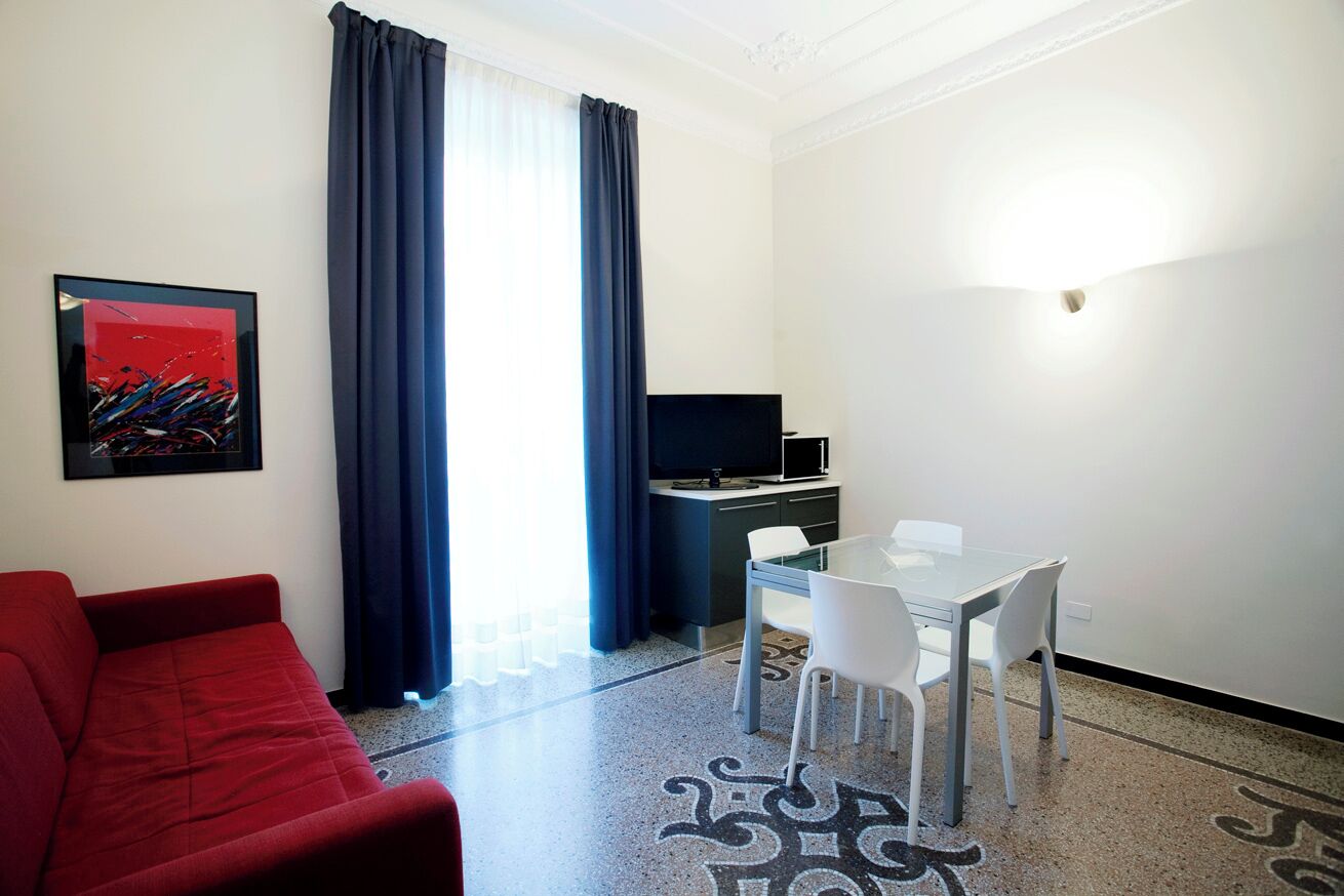 A compact hotel room with a red sofa, a glass dining table with white chairs, a flat-screen TV, and a patterned rug.