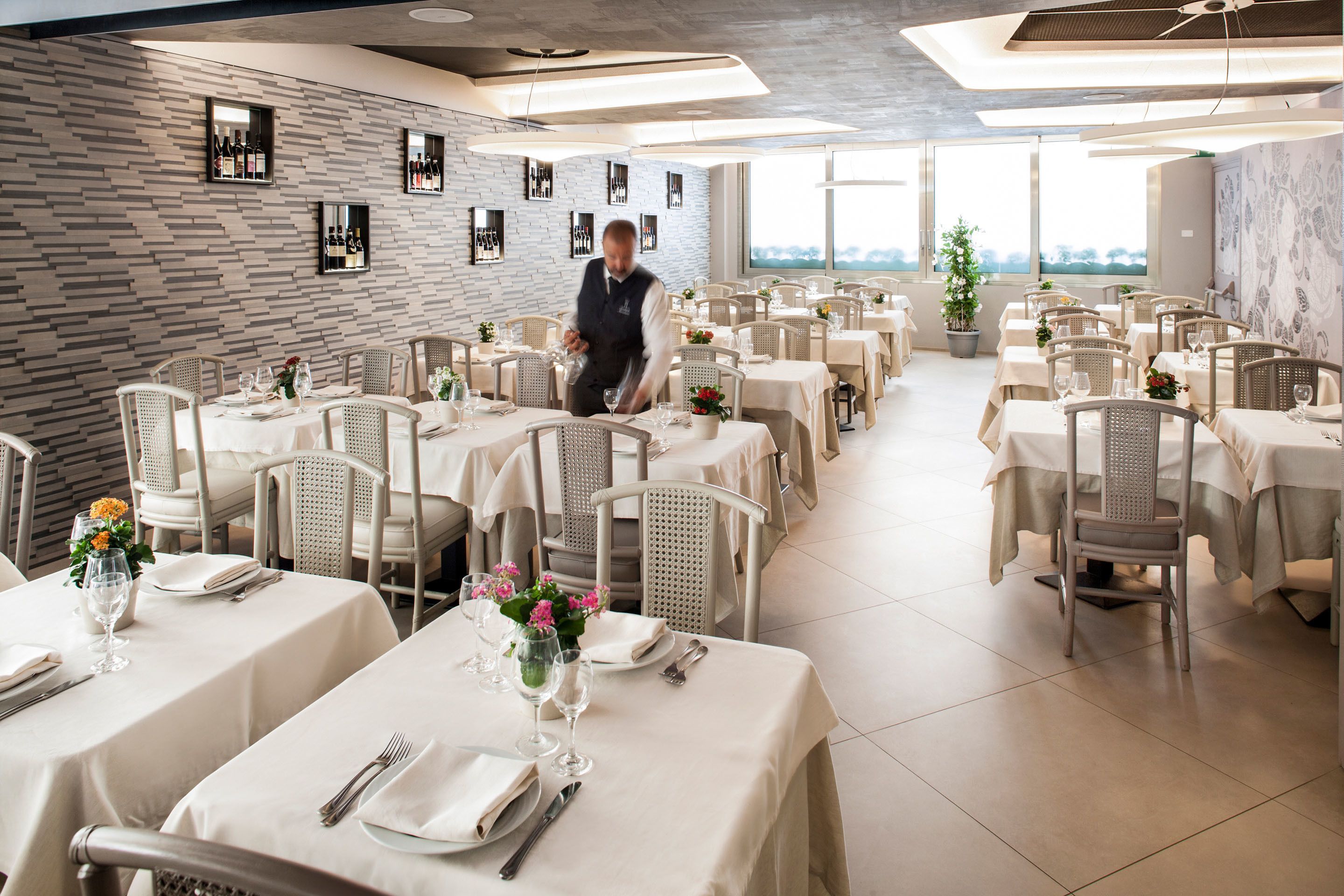 A restaurant with multiple tables set for dining, a waiter attending to guests, and a view of the outdoors through large windows.