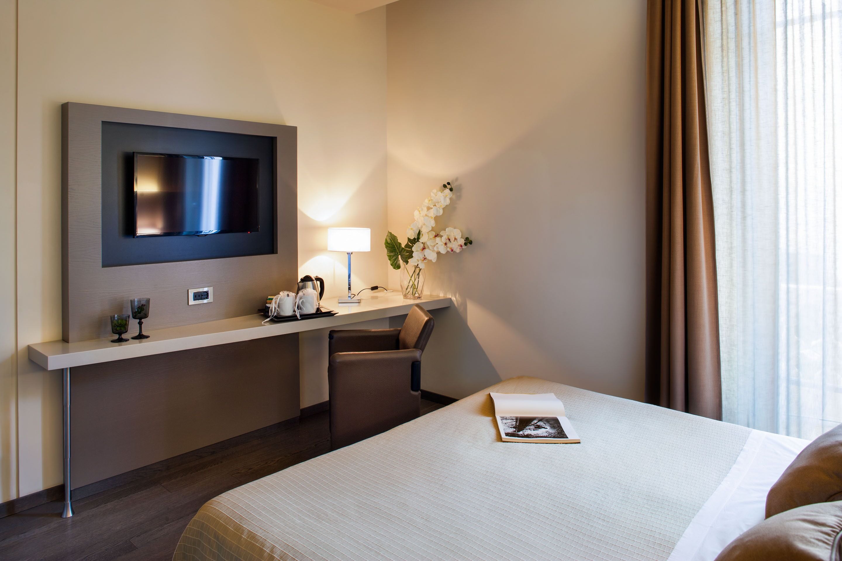Habitación de hotel con cama, escritorio con televisor de pantalla plana, lámpara y un florero con flores.