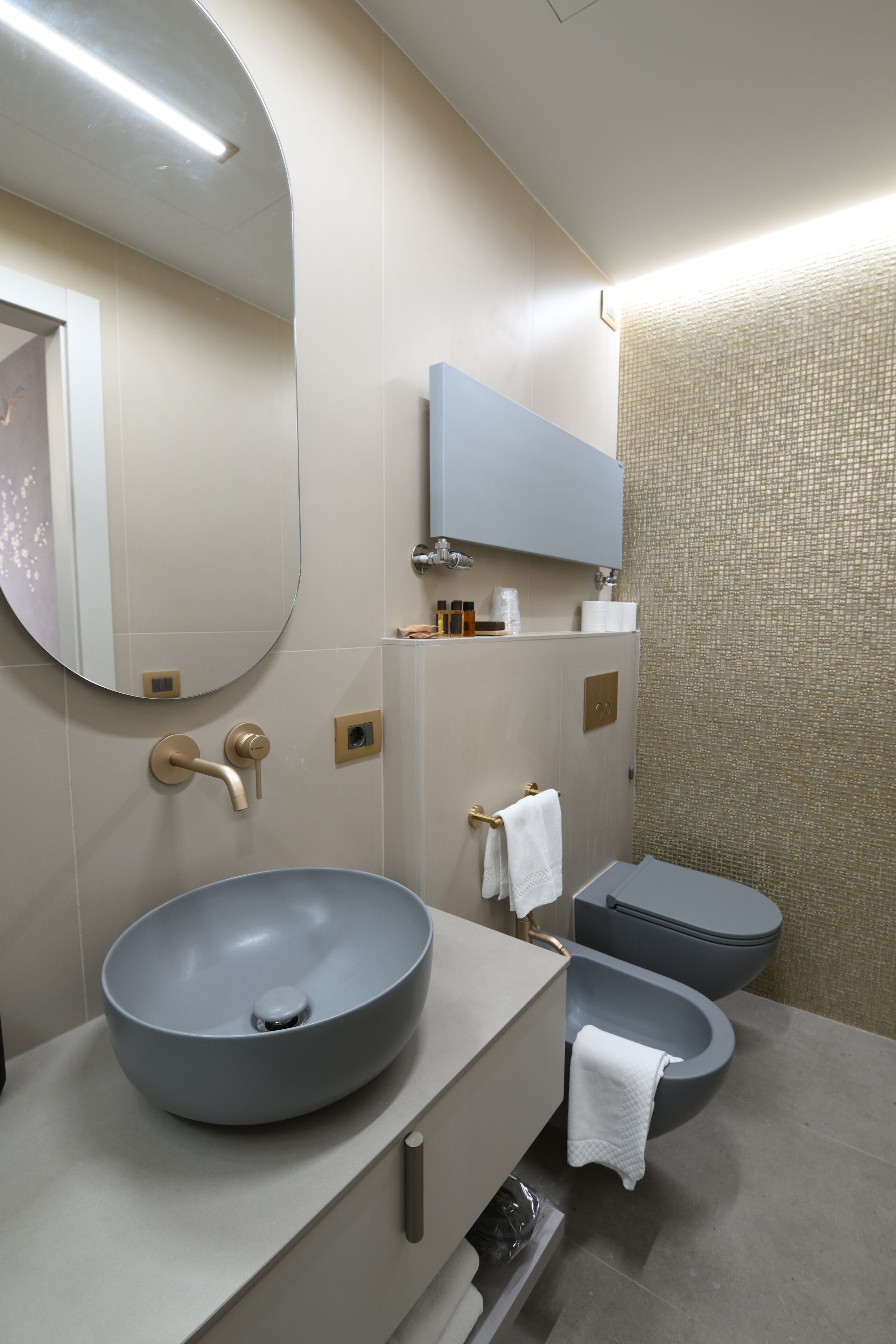 A modern bathroom with a large round sink, a toilet, and a bidet. There is a mirror above the sink and a towel hanging on the wall.