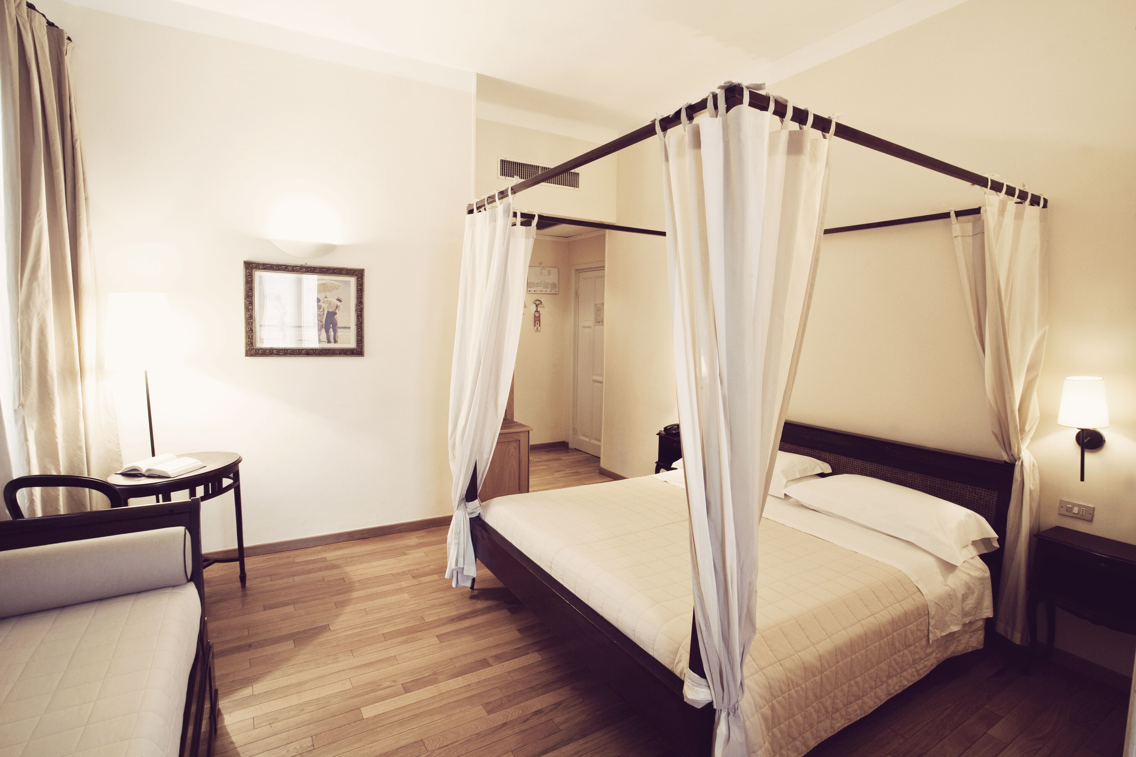 Uma cama de dossel com lençóis brancos, uma mesa de cabeceira com abajur e uma cadeira.