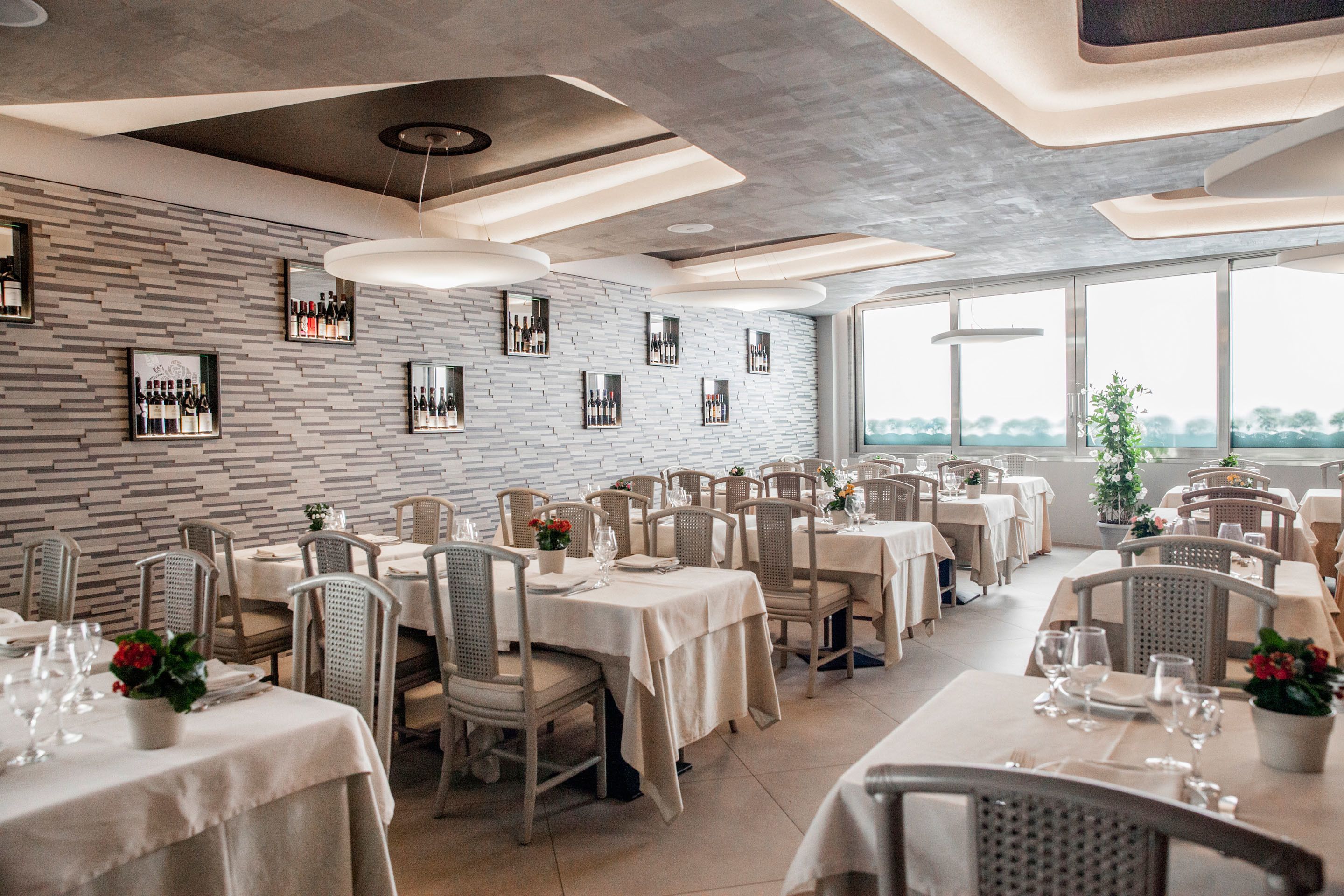 A restaurant with a modern interior, featuring a brick wall with framed wine bottles, white tables with beige tablecloths, and wicker chairs.