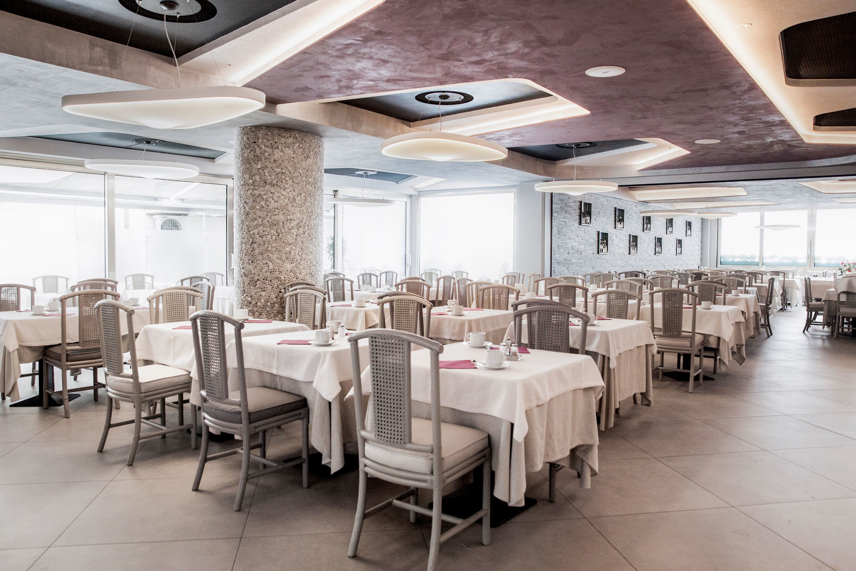 A spacious dining area with multiple tables and chairs, large windows, and a modern ceiling design.