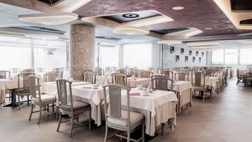 A spacious dining area with multiple tables and chairs, large windows, and a modern ceiling design.