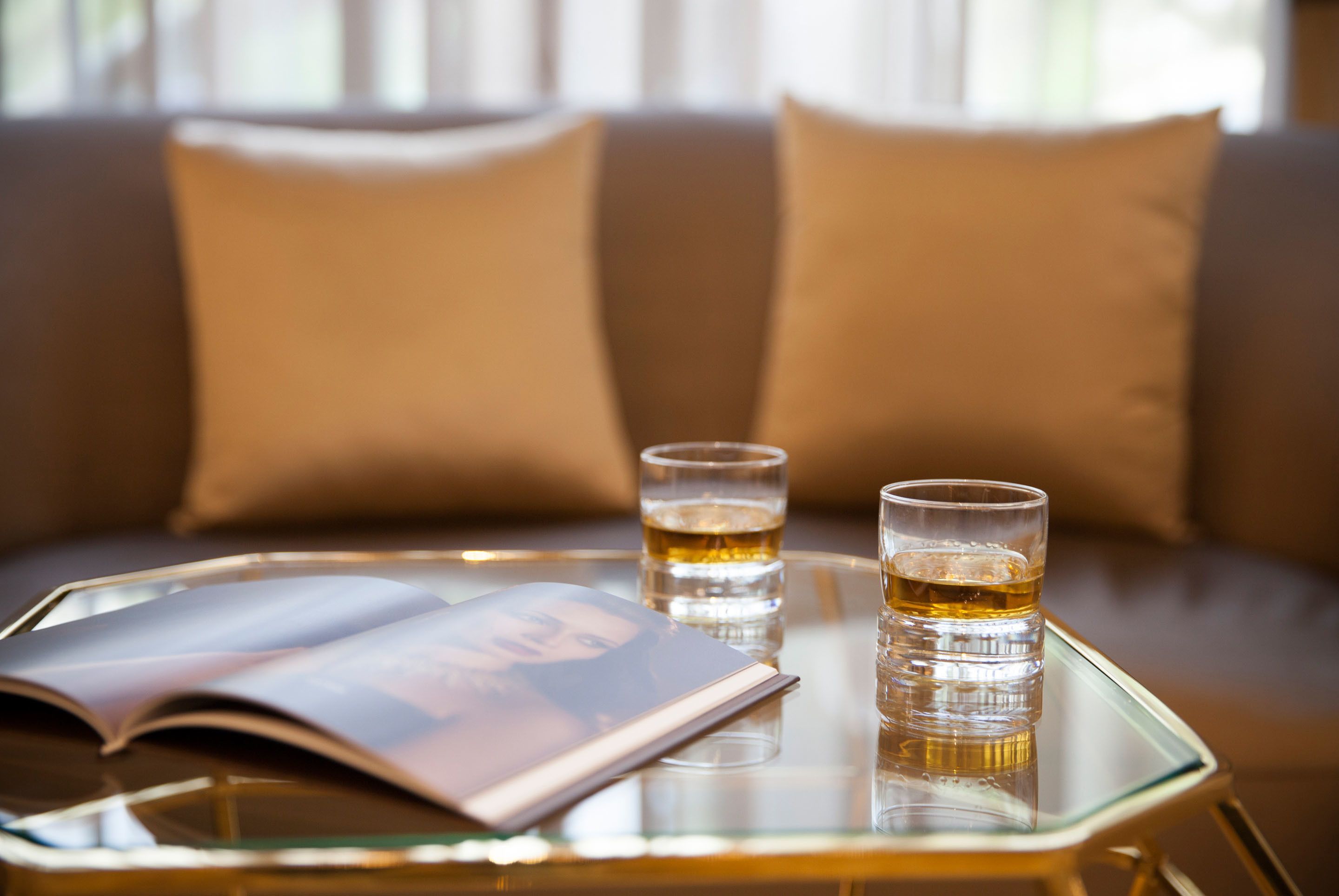Una mesa de vidrio con dos vasos de líquido ámbar, una revista y un álbum de fotos sobre una mesa de vidrio en una habitación con un sofá y almohadas.