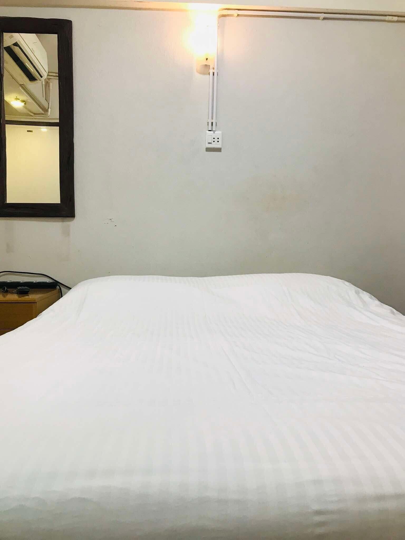 A hotel room with a single bed, a bedside table, a wall-mounted mirror, and a wall socket.