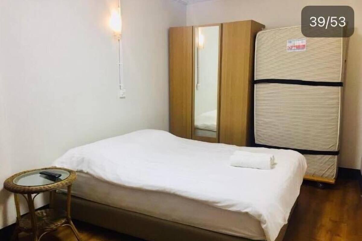 Un lit simple avec un drap blanc, une armoire en bois, une petite table en osier et un matelas muni d’une étiquette.