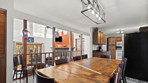 A spacious dining area with a large wooden table and chairs, a modern kitchen in the background, and a view of the outdoors through large windows. - Riley's Retreat  Hot Tub and air conditioning!   (Rapid City)