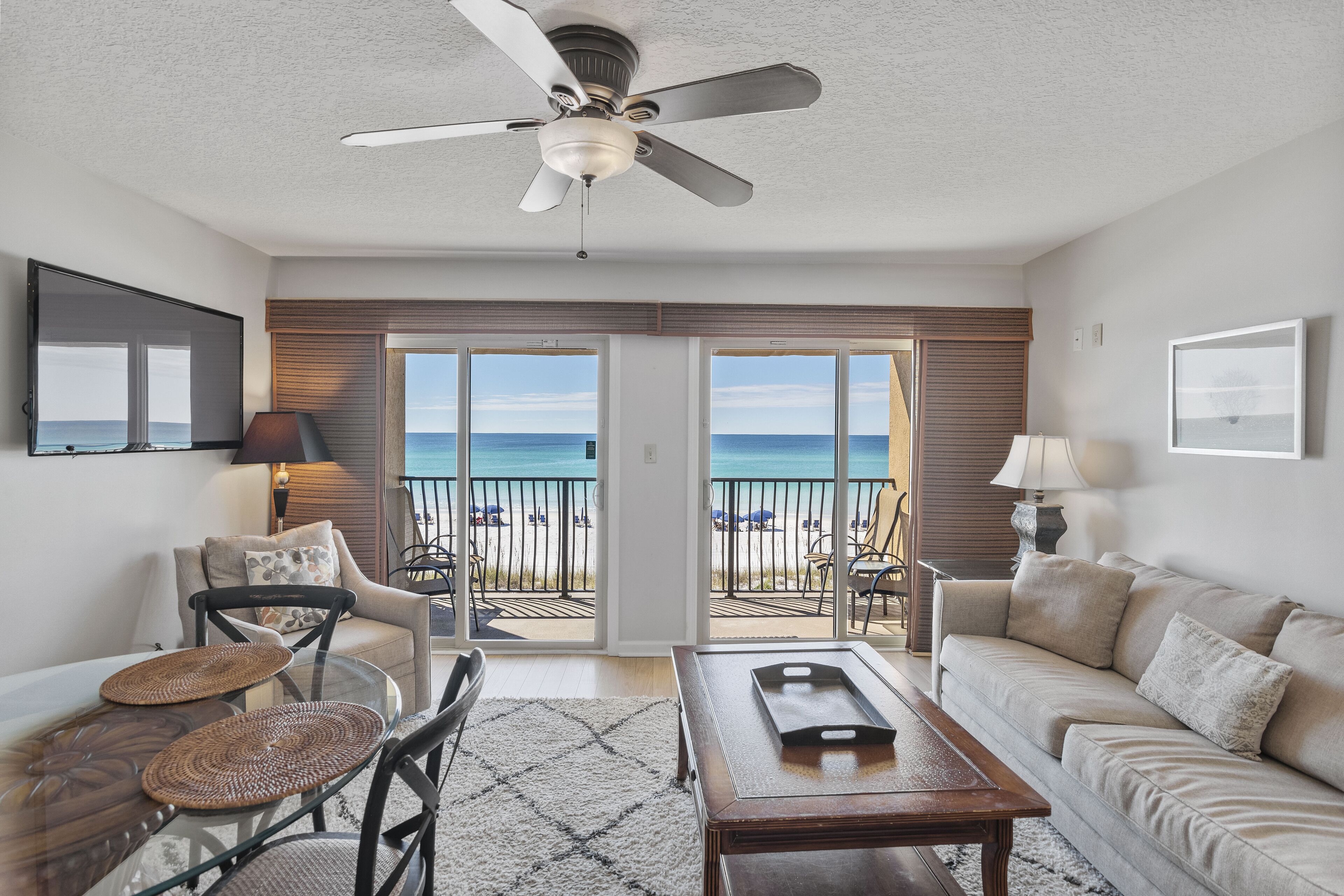 A living room with a glass table, a sofa, and a ceiling fan. There is a view of the beach through large sliding doors.