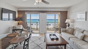 Ein Wohnzimmer mit Glastisch, einer Couch und einem Deckenventilator. Durch große Schiebetüren ist der Blick auf den Strand möglich.