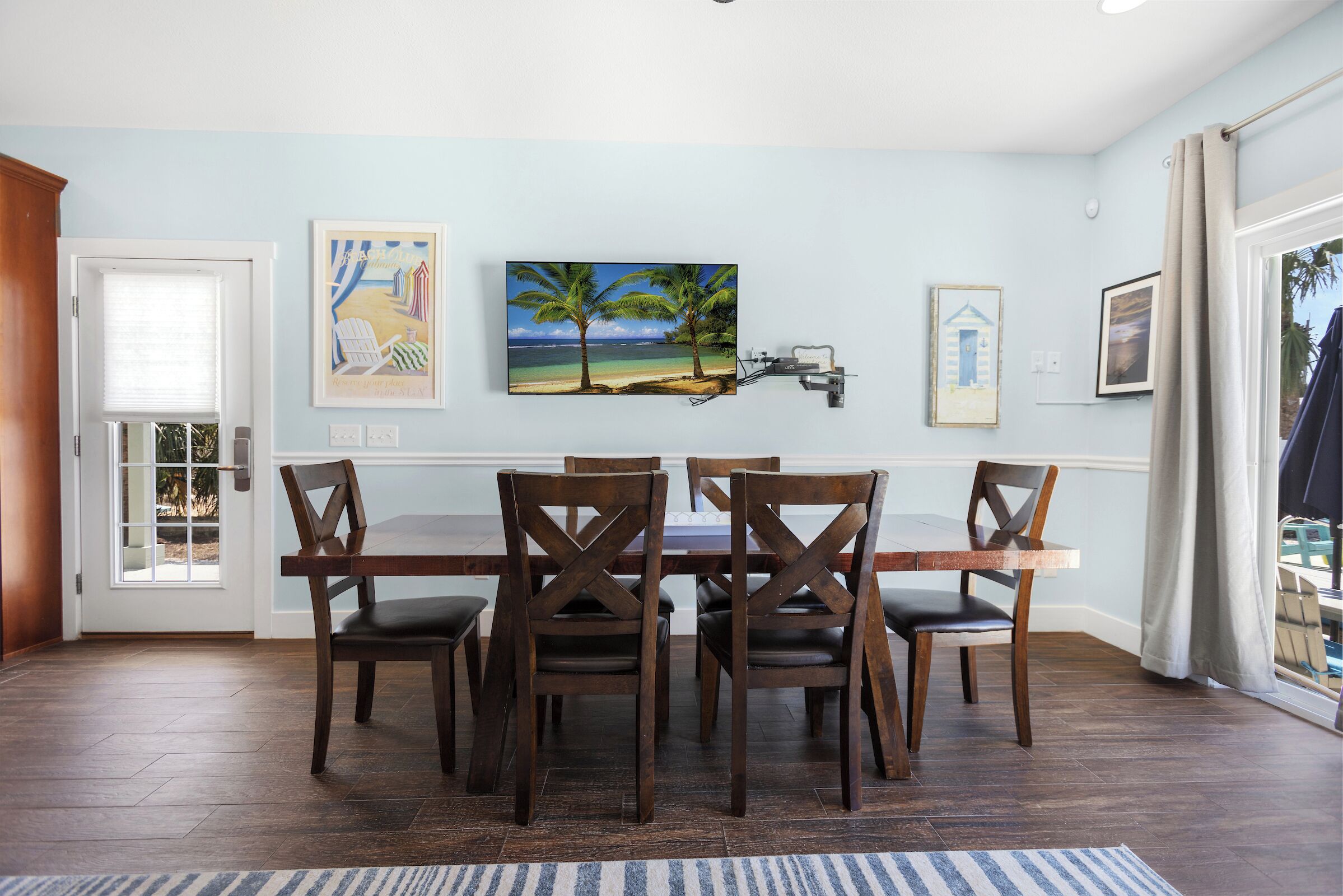 A dining area with a wooden table and chairs, a painting of a beach scene, and a window with curtains.
