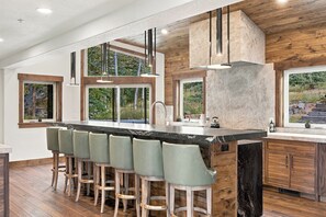 Une cuisine moderne dotée d’un îlot central, de tabourets de bar, de meubles en bois et de grandes fenêtres offrant une vue sur la verdure.