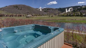 Uma banheira de hidromassagem com vista para um lago, rodeada por um deck de madeira e com montanhas ao fundo.