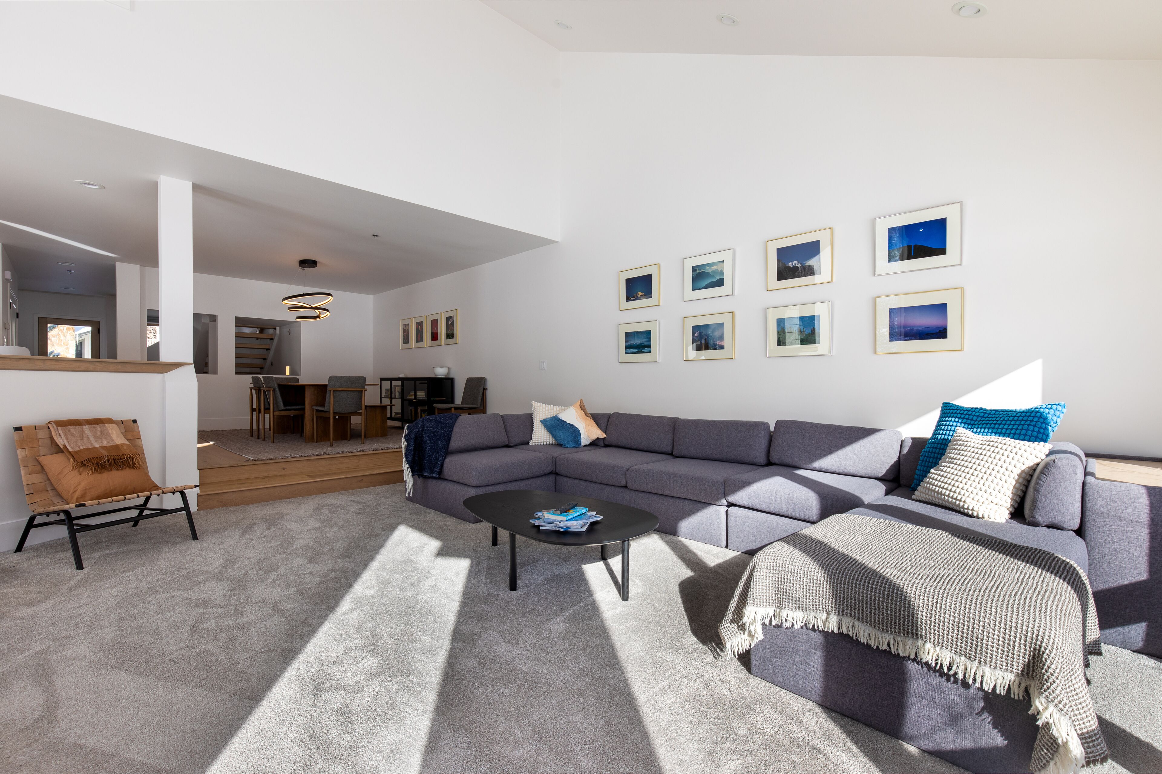 A modern living room with a grey sectional sofa, a coffee table, and a patterned throw blanket. There are framed pictures on the wall and a wooden chair with a throw blanket.