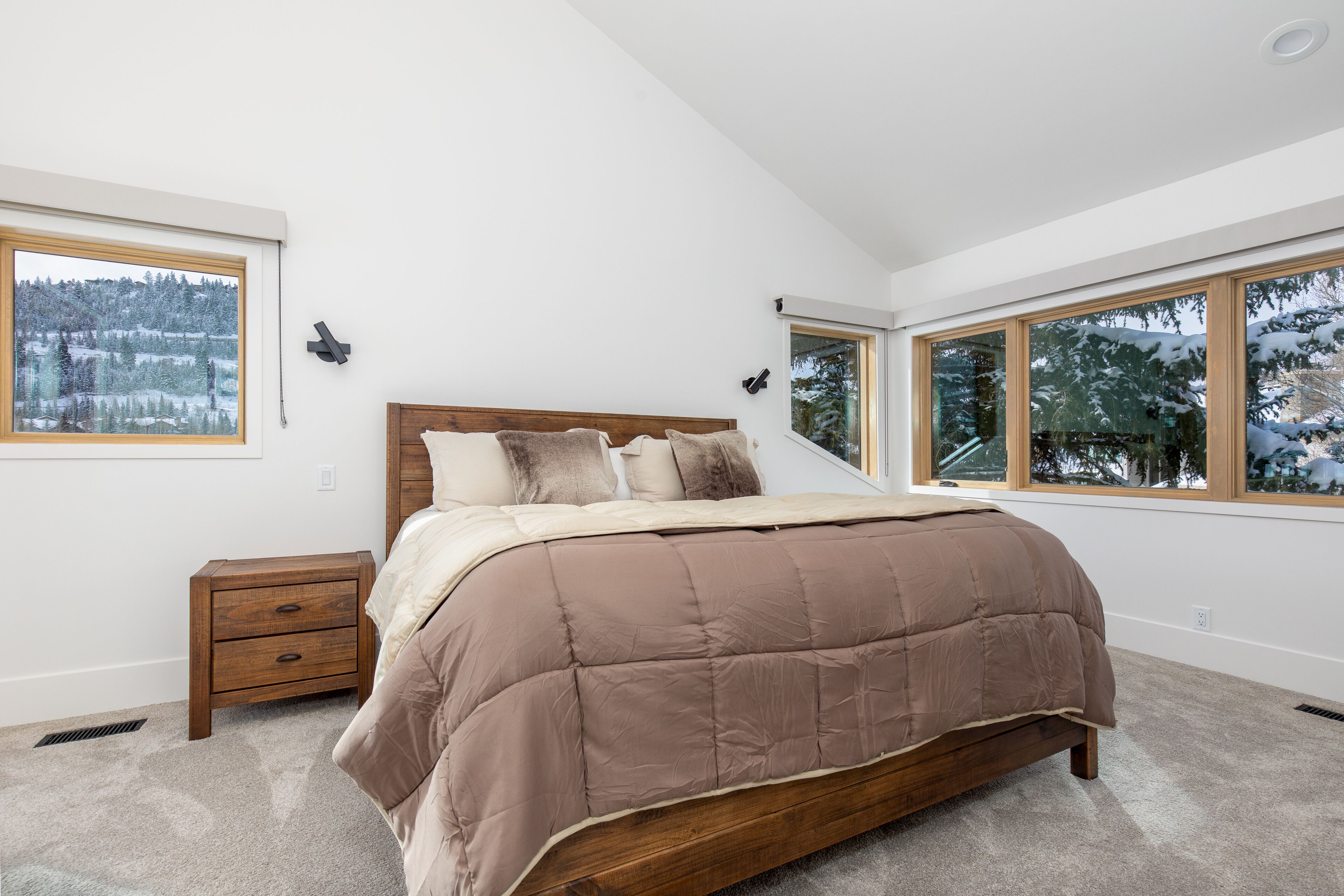 A bedroom with a large bed, wooden headboard, and two bedside tables. There are large windows providing a view of a snowy landscape.