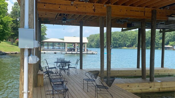 A wooden deck with metal chairs and tables, a ceiling fan, and a view of a lake and buildings in the background. - Watts Bar Lake House (Spring City)