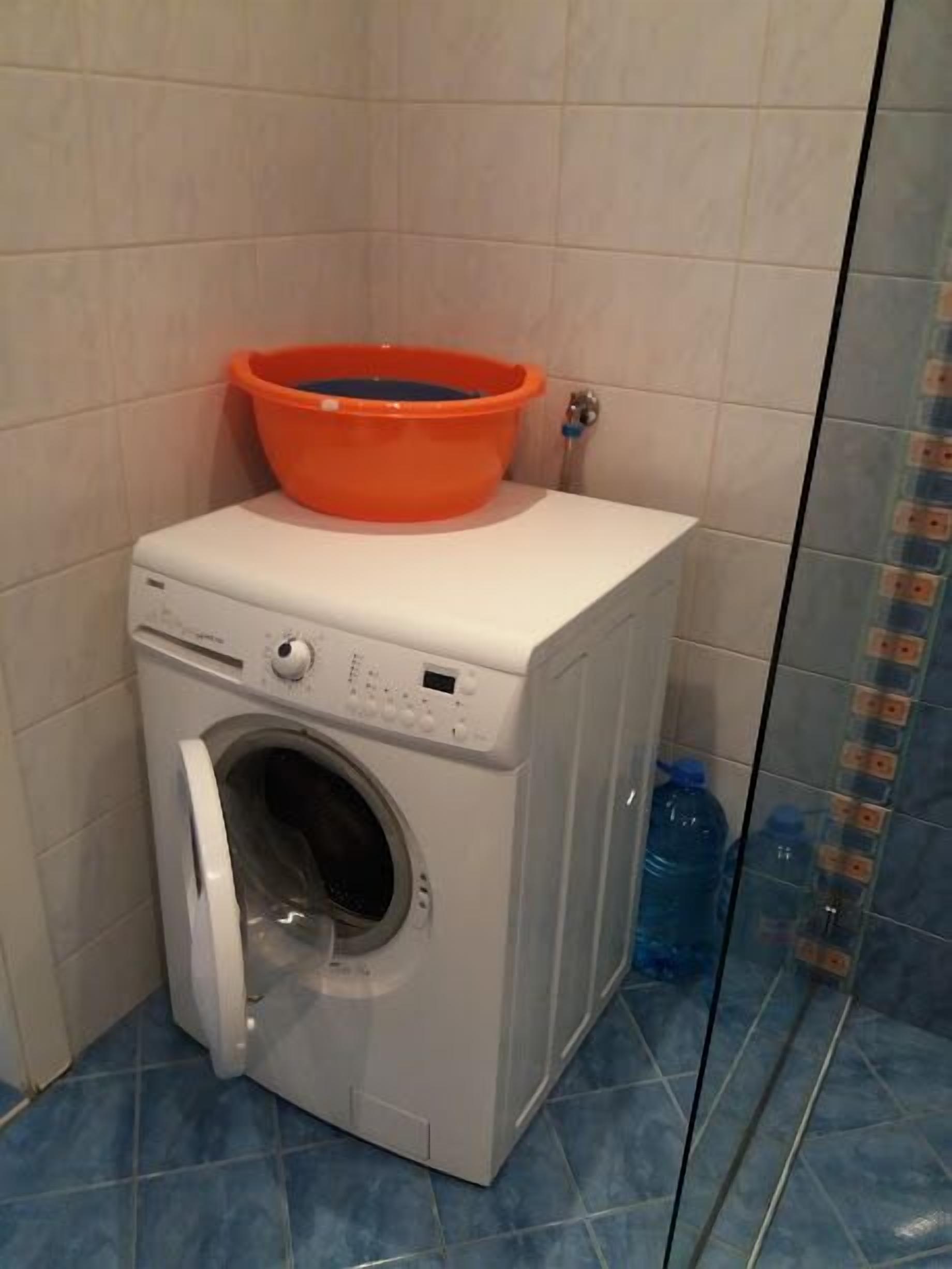 A compact washing machine with a front-loading door, a small orange basin on top, and a blue water bottle beside it.