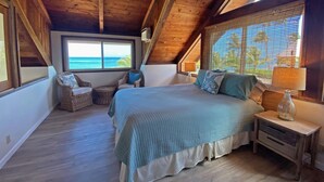 A bedroom with a large bed, two wicker chairs, a nightstand, and a window with a view of the ocean.