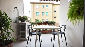 Un pequeño patio al aire libre con una mesa y sillas, rodeado de macetas con plantas y un helecho colgante.