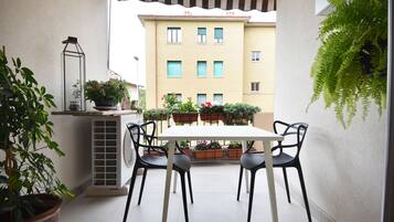 Une petite terrasse extérieure avec une table et des chaises, entourée de plantes en pot et d’une fougère suspendue.