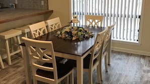 Un coin repas avec une table en bois et des chaises blanches, une cuisine avec des meubles en bois et une grande fenêtre avec des stores.