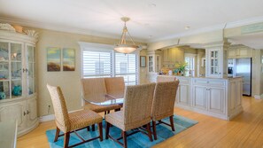 A dining area with a glass-top table and woven chairs, a chandelier, and a view of the kitchen with white cabinetry.