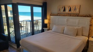 Une chambre d’hôtel avec un grand lit, une table de chevet, une lampe et une vue sur la plage depuis les portes du balcon.