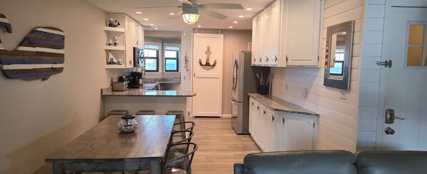 A modern kitchen with white cabinets, a wooden dining table, and a view of the ocean through the windows.