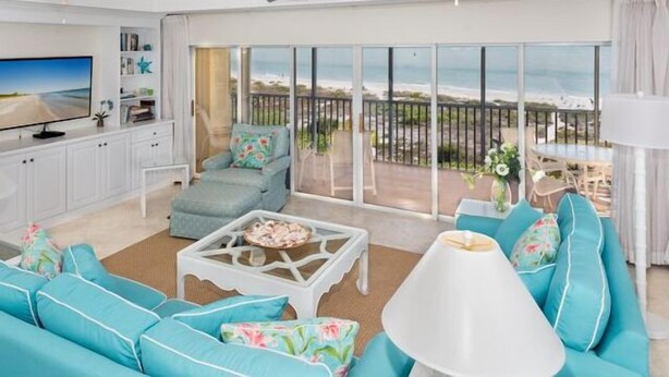 En moderne stue med et stort fjernsyn, turkisfarvede sofaer, et sofabord og en balkon med udsigt til stranden.