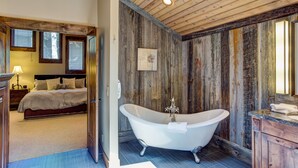 Une salle de bain rustique avec une baignoire sur pieds, des murs en bois et un lit visible dans la pièce adjacente.