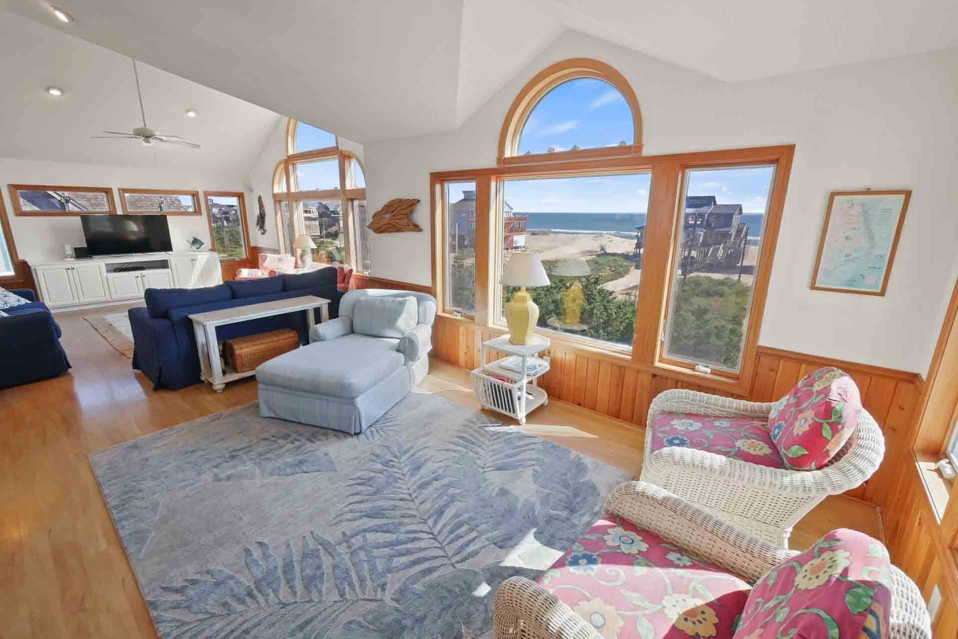 A spacious living room with a large window offering a view of the beach, a ceiling fan, and a television on a cabinet.