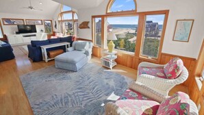A spacious living room with a large window offering a view of the beach, a ceiling fan, and a television on a cabinet. - Semi-Oceanfront Dog Friendly Avon Home w/ Private Pool, Hot Tub, and Game Room (Avon)