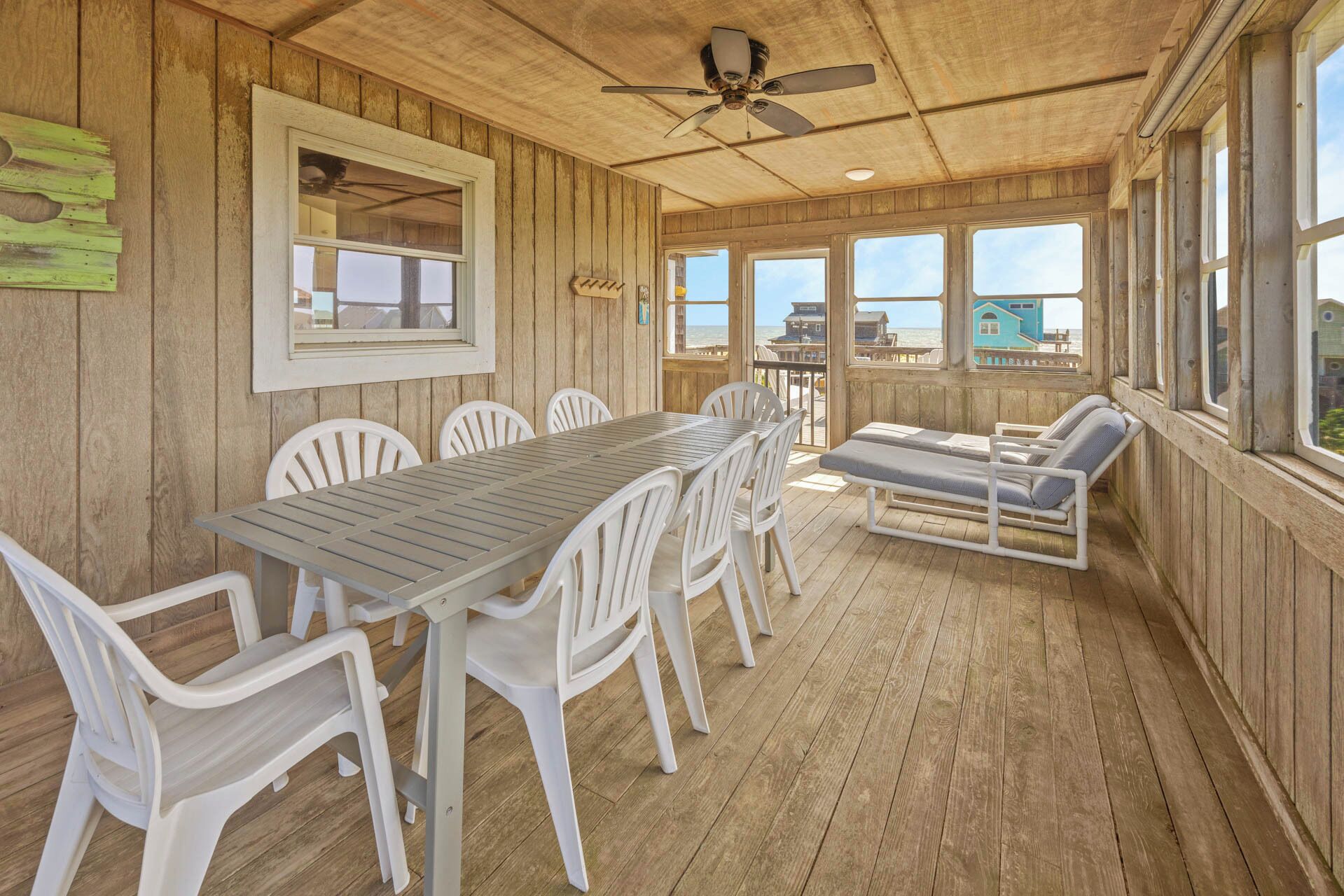 A wooden deck with a table and chairs, a ceiling fan, and large windows overlooking the beach.