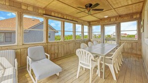 A covered patio with a wooden floor, white chairs, a table, and large windows offering views of houses and greenery. - Semi-Oceanfront Dog Friendly Avon Home w/ Private Pool, Hot Tub, and Game Room (Avon)