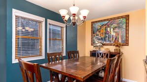 A dining room with a wooden table and chairs, two windows with blinds, a chandelier, and a painting on the wall. - Heart of PC! 2 blocks from lift! Shuttle to Main in 3 minutes! Cozy w/ hot tub! (Park City)
