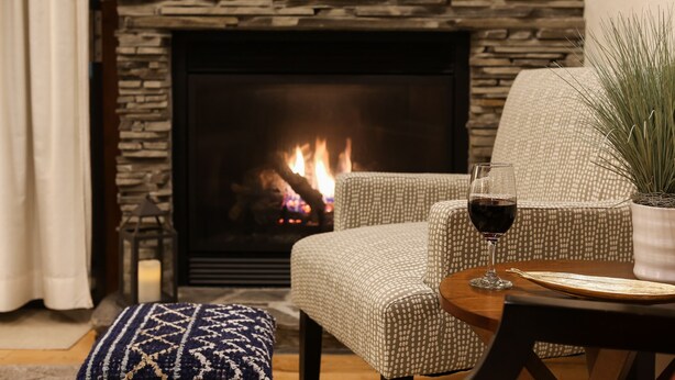 Un salon chaleureux avec une cheminée en pierre, un fauteuil à motifs, une table basse en bois et un verre de vin rouge.